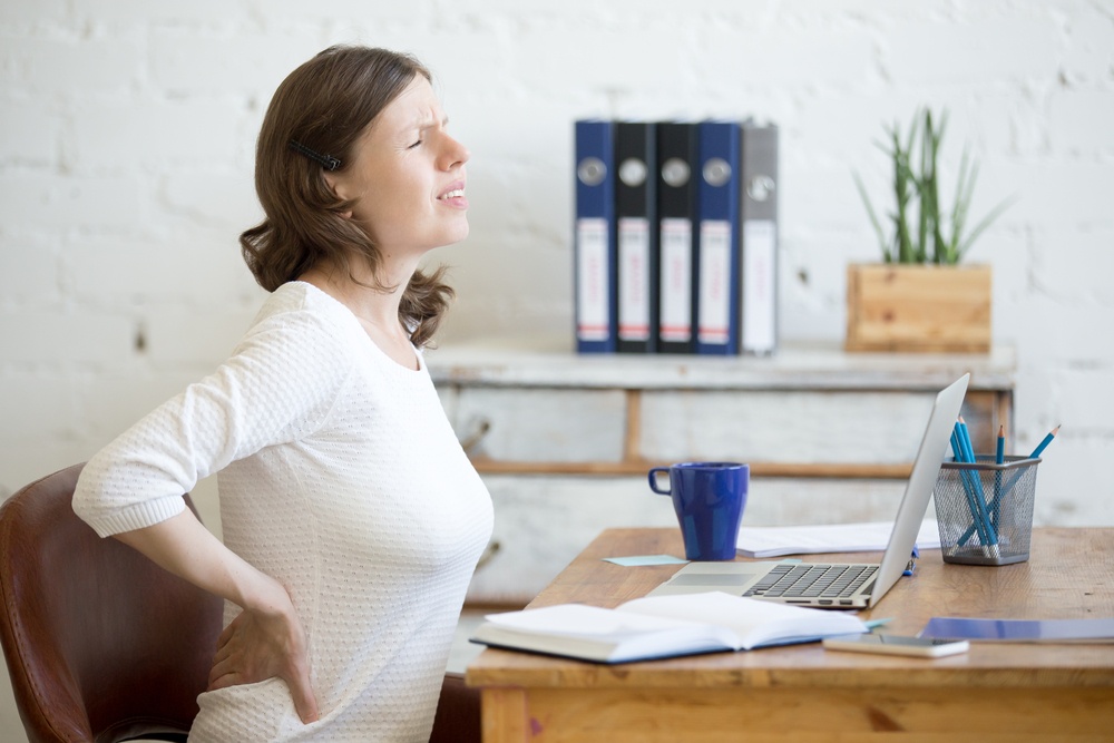 Young stressed businesswoman sitting in front of laptop and holding her waist with pained expression. Business woman feeling pain, touching aching back, suffering from backache after working on pc