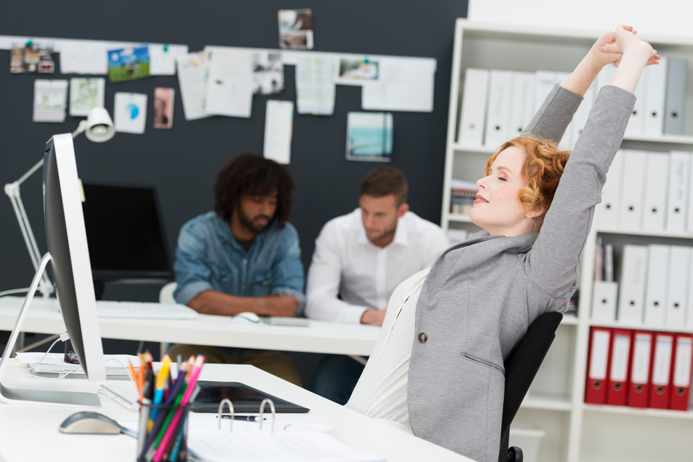 Happy beautiful contented young businesswoman relaxing in her chair stretching her arms in the air in a busy office with multiethnic male business partners